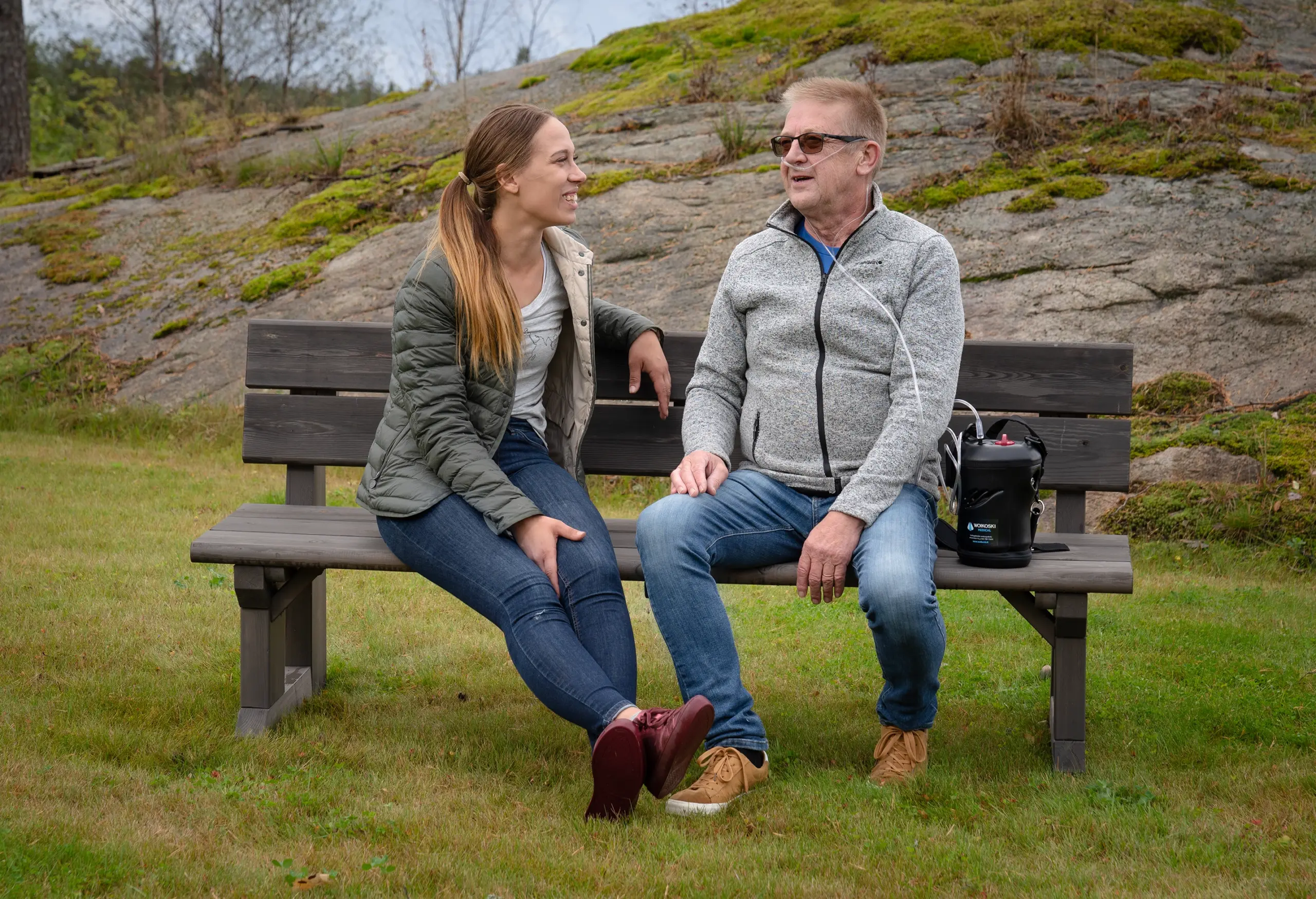 Nuori nainen ja vanhempi mies istuvat puiston penkillä. Miehellä on happiviikset ja kannettava nestehappilaite käytössä.